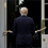 President Trump stands outside a White House entrance, unable to enter due to the stick he carries being longer than the width of the doorway.